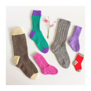 May include: A collection of colorful, hand-knitted socks in various sizes and designs. The socks feature a range of colors, including brown, green, purple, pink, gray, and red. Some socks have contrasting cuffs, heels, and toes. The arrangement is on a white surface.