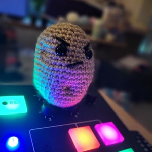 May include: A crocheted brown potato with angry black eyes and a frown, sitting on top of a black music mixer with colorful lights.