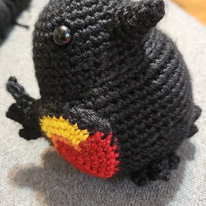 May include: A black crocheted bird with red, yellow, and orange accents on its chest. The bird has black eyes and is sitting on a gray surface.