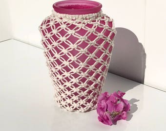 May include: A pink glass vase with a white crocheted cover. The vase is sitting on a white surface with a small cluster of pink flowers next to it.