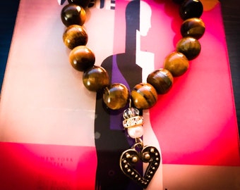 May include: A brown tiger's eye beaded bracelet with a gold heart charm and a white pearl. The bracelet is on a pink and white background with the text "NEW YORK CITY".