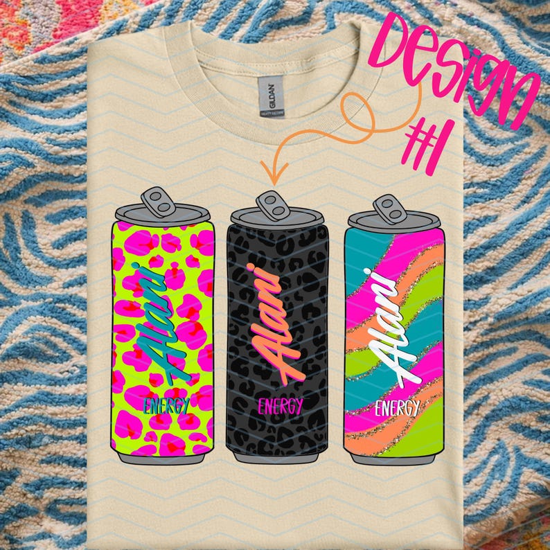 May include: A cream-colored t-shirt featuring three stylized energy drink cans. Each can has a different design: neon pink leopard print, black leopard print, and a teal and pink striped design. The word "Alani" is written vertically on each can, with the word "Energy" below.