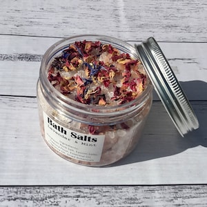 Op de afbeelding: Een glazen pot gevuld met badzout, gedroogde lavendel en rozenblaadjes. Het etiket vermeldt "Bath Salts Lavender & Mint" en somt de ingredi&euml;nten op: Himalaya-rotszout, Epsom-zouten, tapiocameel, etherische olie en gedroogde kruiden.