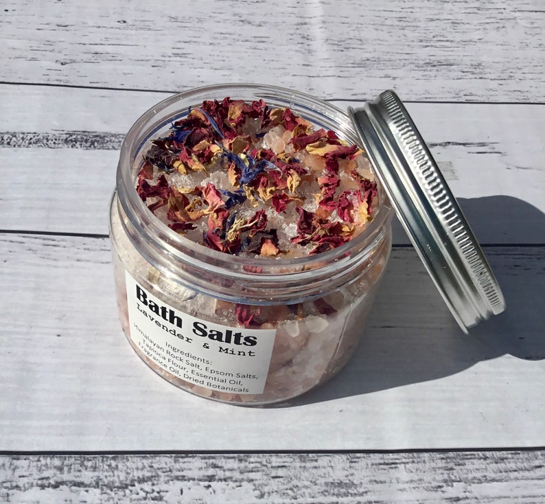Op de afbeelding: Een glazen pot gevuld met badzout, gedroogde lavendel en rozenblaadjes. Het etiket vermeldt "Bath Salts Lavender & Mint" en somt de ingredi&euml;nten op: Himalaya-rotszout, Epsom-zouten, tapiocameel, etherische olie en gedroogde kruiden.