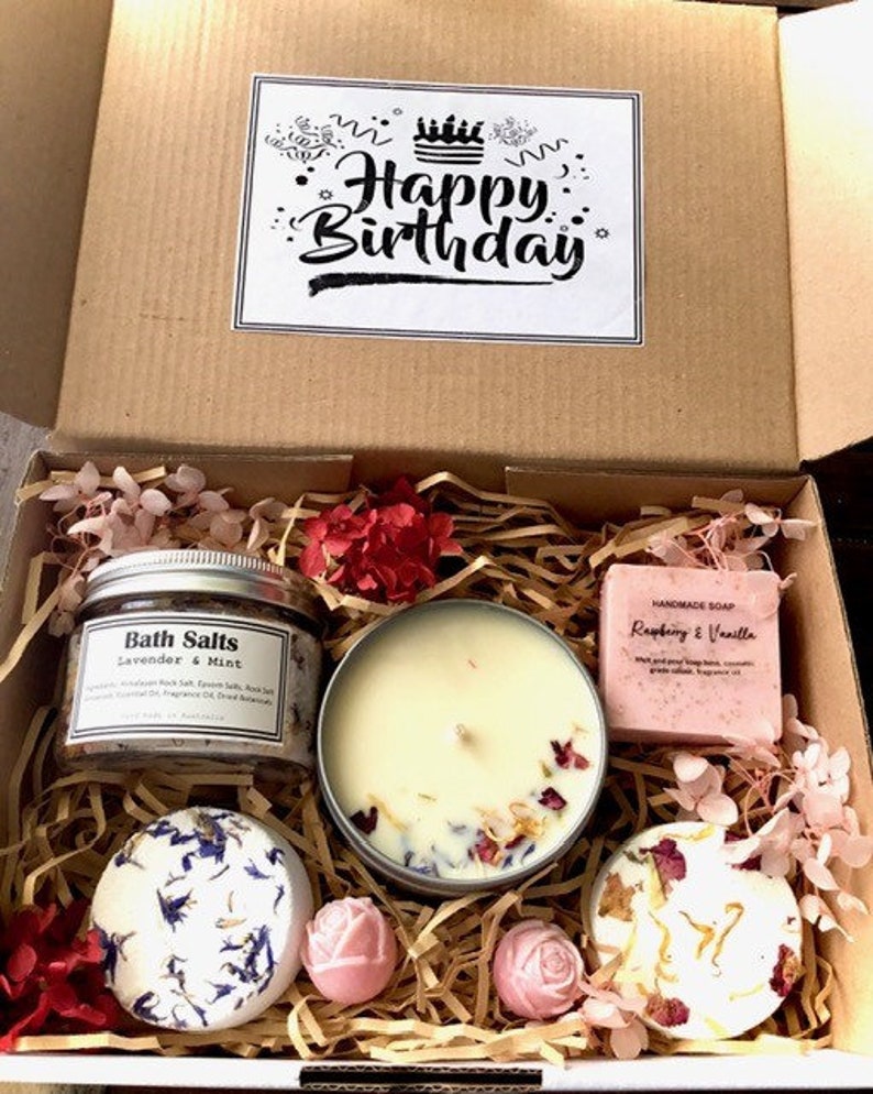 Op de afbeelding: Een cadeaudoos gevuld met bad- en lichaamsverzorgingsproducten. De doos bevat een kaars, badzout, badbommen en zeep. Het badzout is gelabeld met "Bath Salts Lavender & Mint". De zeep is gelabeld met "Handmade Soap Raspberry & Vanilla". De doos is versierd met een kaartje met "Happy Birthday".