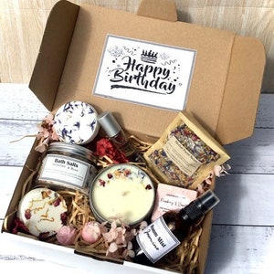 Op de afbeelding: Een witte cadeaudoos gevuld met verschillende bad- en lichaamsverzorgingsproducten, waaronder badzout, een kaars, een zeep en een kamerspray. De doos is versierd met een kaart "Happy Birthday" en roze bloemen.