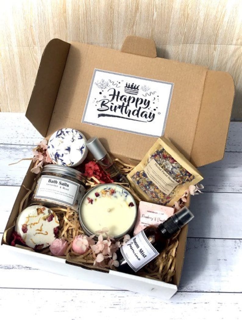 Op de afbeelding: Een witte cadeaudoos gevuld met verschillende bad- en lichaamsverzorgingsproducten, waaronder badzout, een kaars, een zeep en een kamerspray. De doos is versierd met een kaart "Happy Birthday" en roze bloemen.