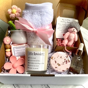 May include: A self-care gift box containing a white towel with a pink ribbon, a candle with text, a pink teddy bear, a bath bomb, body oil, and soap. The box includes pink and white items, such as a flower-shaped item and a small pot.