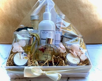 May include: A gift basket filled with bath and body products, including a bottle of body oil, a bottle of hand soap, a jar of bath salts, a jar of lip balm, and two candles. The basket is decorated with white flowers and a yellow ribbon.