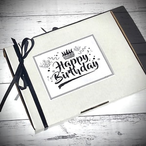 Op de afbeelding: Een witte cadeaudoos met een zwart lint eromheen gebonden. De doos heeft een sticker met de tekst "Happy Birthday" met een taart- en confetti-ontwerp.