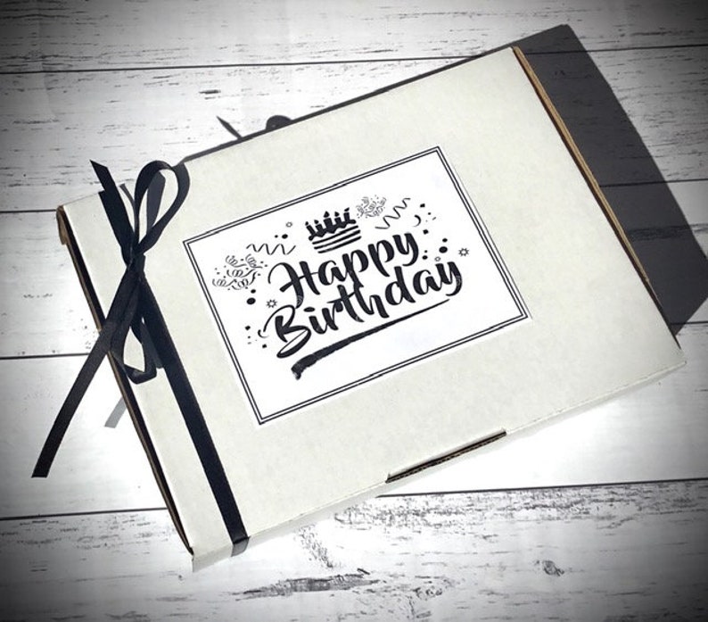 Op de afbeelding: Een witte cadeaudoos met een zwart lint eromheen gebonden. De doos heeft een sticker met de tekst "Happy Birthday" met een taart- en confetti-ontwerp.