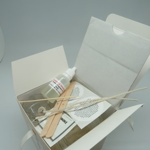 May include: A white box with a clear glass jar, two wooden sticks, a small bottle of liquid, and white wicks. The box is open and the contents are visible. The jar has a label that says "CANDLE" and "Made in Australia". The bottle has a label that says "Soy Wax & Soy Fragrance Oil".