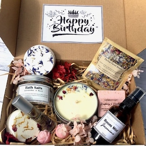 Op de afbeelding: Een cadeaudoos gevuld met bad- en lichaamsverzorgingsproducten, waaronder badzout, een kaars, zeep, een gezichtsstoom en een kamernevel. De doos is versierd met roze en rode bloemen en een kaart met de tekst "Happy Birthday".