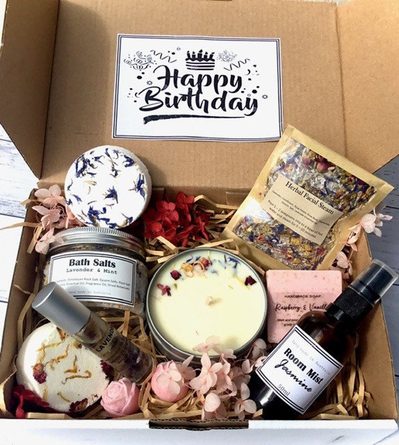 Op de afbeelding: Een cadeaudoos gevuld met bad- en lichaamsverzorgingsproducten, waaronder badzout, een kaars, zeep, een gezichtsstoom en een kamernevel. De doos is versierd met roze en rode bloemen en een kaart met de tekst "Happy Birthday".