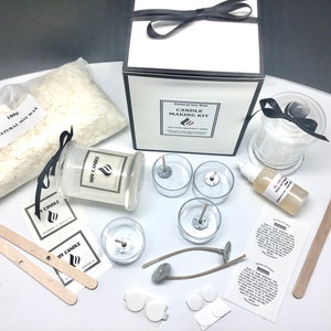 May include: Candle making kit with soy wax flakes, glass jars, wicks, and labels. The kit includes instructions and a bottle of fragrance oil. The kit is packaged in a white box with a black ribbon.