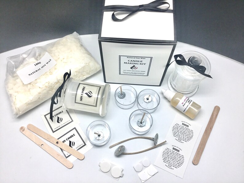 May include: Candle making kit with soy wax flakes, glass jars, wicks, and labels. The kit includes instructions and a bottle of fragrance oil. The kit is packaged in a white box with a black ribbon.