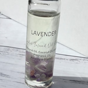 Op de afbeelding: Een heldere glazen rollerfles met een witte dop gevuld met een lavendelgeurende oliemengsel. De fles bevat amethistkristallen en een etiket met de tekst "LAVENDER Crystal Infused Oil Roll Almond Oil, Coconut Oil, Essence Fragrance Oil, Amethyst Crystals".