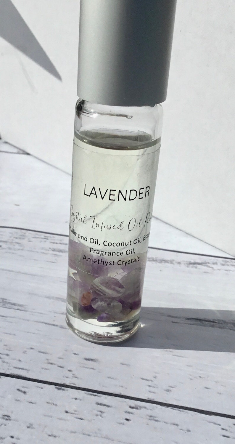 Op de afbeelding: Een heldere glazen rollerfles met een witte dop gevuld met een lavendelgeurende oliemengsel. De fles bevat amethistkristallen en een etiket met de tekst "LAVENDER Crystal Infused Oil Roll Almond Oil, Coconut Oil, Essence Fragrance Oil, Amethyst Crystals".
