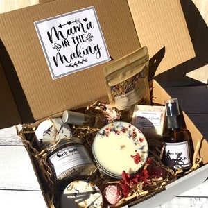 May include: A gift box labelled "Mama in the Making" contains a candle, bath salts, soap, and other self-care items. The candle is decorated with dried flowers. The box is filled with straw and other decorative elements.
