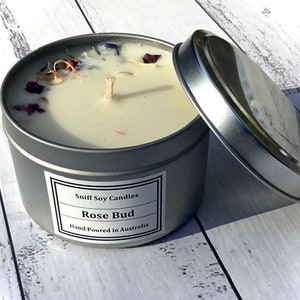 Op de afbeelding: Een zilveren blikje met daarin een witte sojawas-kaars. De kaars is versierd met gedroogde bloemen en heeft een etiket met de tekst "Sniff Soy Candles Rose Bud Hand Poured in Australia".