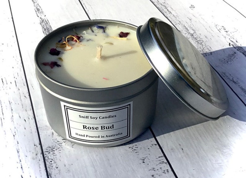 Op de afbeelding: Een zilveren blikje met daarin een witte sojawas-kaars. De kaars is versierd met gedroogde bloemen en heeft een etiket met de tekst "Sniff Soy Candles Rose Bud Hand Poured in Australia".