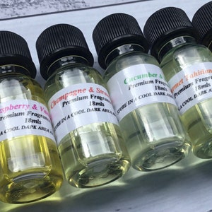 May include: A row of six small glass bottles with black caps, each containing a clear liquid. The bottles are labelled with different scents, including Raspberry & Vanilla, Champagne & Strawberry, Cucumber & Mint, Gourmet Tahitian Vanilla, and Raspberry. Each label includes the text "Premium Fragrance Oil" and "18mls". The bottles are arranged on a white wooden surface.