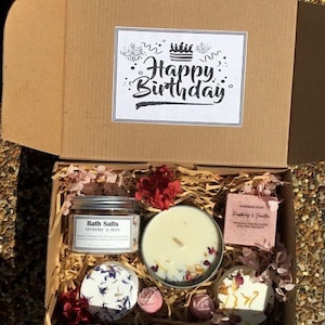 Op de afbeelding: Een bruine kartonnen cadeaudoos met een witte kaart waarop "Happy Birthday" staat. De doos is gevuld met verschillende bad- en lichaamsverzorgingsproducten, waaronder badzout, kaarsen en zeep. De producten zijn decoratief gerangschikt met roze en rode bloemen.