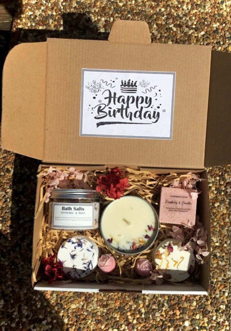 Op de afbeelding: Een bruine kartonnen cadeaudoos met een witte kaart waarop "Happy Birthday" staat. De doos is gevuld met verschillende bad- en lichaamsverzorgingsproducten, waaronder badzout, kaarsen en zeep. De producten zijn decoratief gerangschikt met roze en rode bloemen.