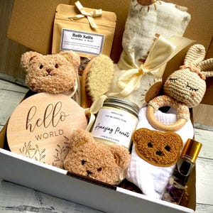 May include: A baby gift box containing various items. It features a wooden sign with "hello world" text, a teddy bear, bath salts, a brush, a candle, a bib, a wooden teether, a bunny, and a bottle of oil.