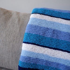 May include: A striped knitted throw blanket in shades of blue, white, and navy blue. The blanket is folded and draped over a light gray couch.