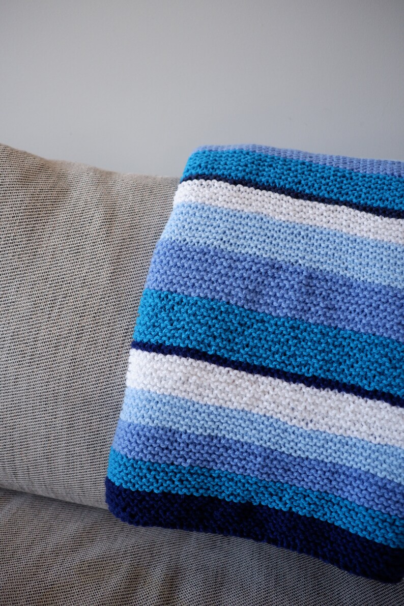 May include: A striped knitted throw blanket in shades of blue, white, and navy blue. The blanket is folded and draped over a light gray couch.