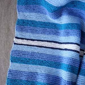 May include: A blue and white striped knitted blanket with a textured pattern. The blanket is folded and draped over a couch.