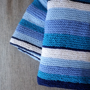 May include: A blue and white striped knitted blanket with a textured pattern. The blanket is folded in half and is lying on a gray surface.