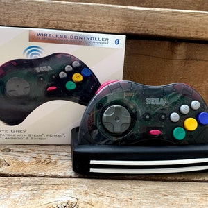 May include: A black and grey Sega Saturn wireless controller with a translucent grey shell. The controller is compatible with Steam, PC/Mac, PS3, Android, and Switch. The box says "SEGA SATURN" and "WIRELESS CONTROLLER".