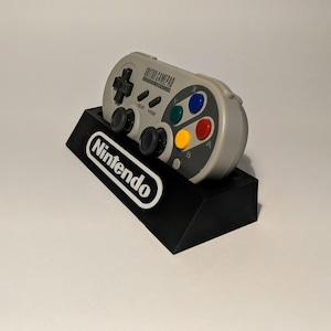 May include: A gray retro gamepad with colored buttons, resting on a black stand with the Nintendo logo. The gamepad has a directional pad, start and select buttons, and four colored action buttons.