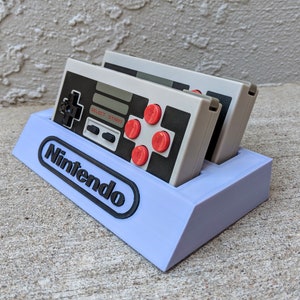 May include: A purple plastic stand with the word "Nintendo" printed on the front. Two gray Nintendo Entertainment System controllers are sitting in the stand.