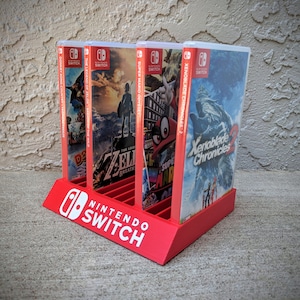 May include: Red plastic stand for Nintendo Switch game cartridges. The stand holds six game cartridges and has the Nintendo Switch logo printed on the front. The stand is filled with game cartridges for games like Super Mario Odyssey, The Legend of Zelda: Breath of the Wild, and Xenoblade Chronicles 2.