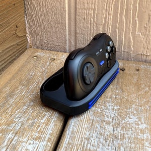 May include: A black Sega Genesis controller with a blue and black charging dock. The controller has a six-button layout and a directional pad.