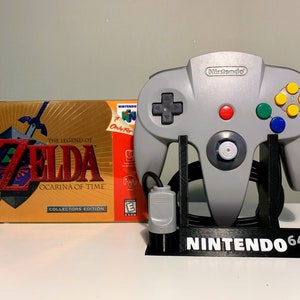 May include: A gray Nintendo 64 controller with a black stand and a black and gray controller cord. The controller is on a white surface. The box for the game "The Legend of Zelda: Ocarina of Time" is in the background. The box is gold with a red and black design.