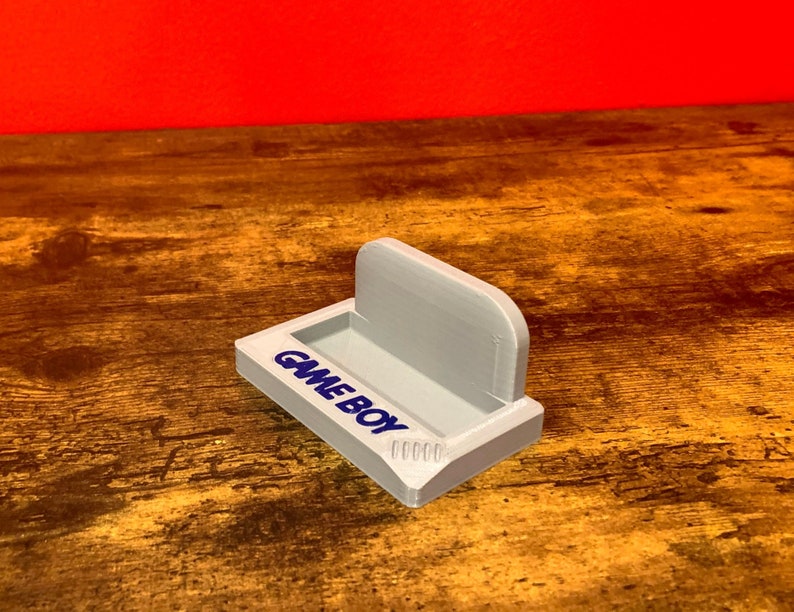 May include: A white plastic stand with the words "GAME BOY" printed on the front. The stand is designed to hold a Game Boy handheld gaming device.