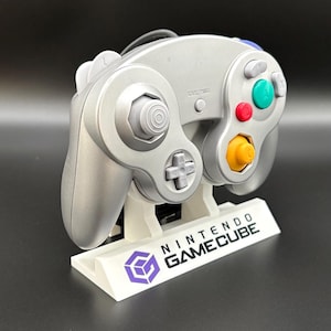 Peut inclure: Manette Nintendo GameCube argentée sur un support blanc avec le logo et le texte GameCube. La manette possède des boutons colorés et un pavé directionnel. Le support porte l'inscription "NINTENDO GAMECUBE".