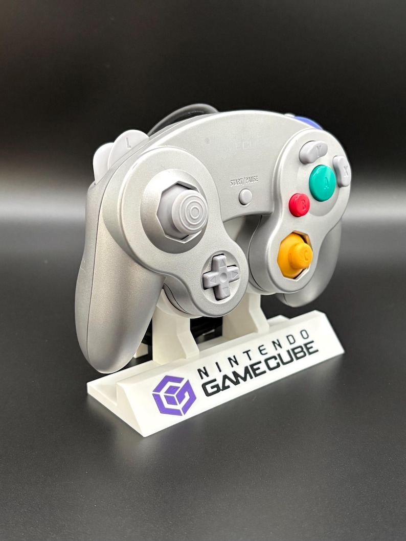 May include: Silver Nintendo GameCube controller on a white stand with the GameCube logo and text. The controller has colored buttons and a directional pad. The stand has the words "NINTENDO GAMECUBE" printed on it.
