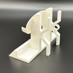 May include: A white 3D-printed phone stand with a unique, geometric design. The stand features a base, a back support, and a series of angled and curved elements. The stand is designed to hold a phone in a vertical position.