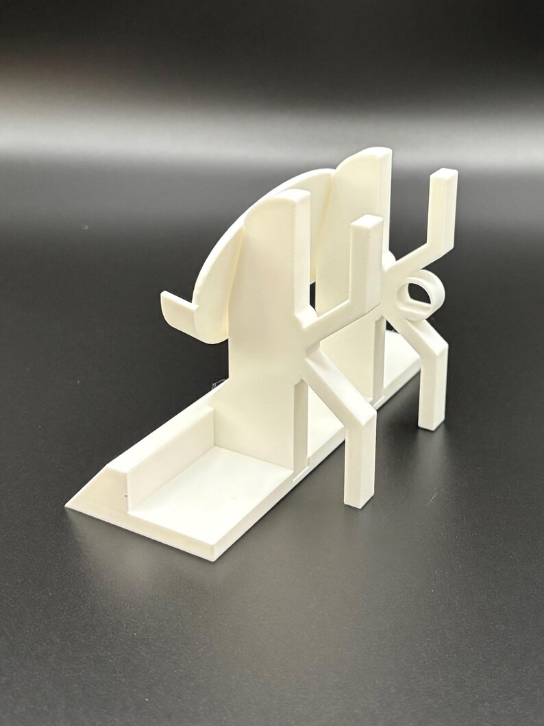 May include: A white 3D-printed phone stand with a unique, geometric design. The stand features a base, a back support, and a series of angled and curved elements. The stand is designed to hold a phone in a vertical position.