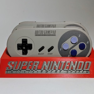 May include: A red plastic stand with the words "SUPER NINTENDO ENTERTAINMENT SYSTEM" printed on the front. Two gray 8BitDo Gamepad controllers are sitting on the stand.