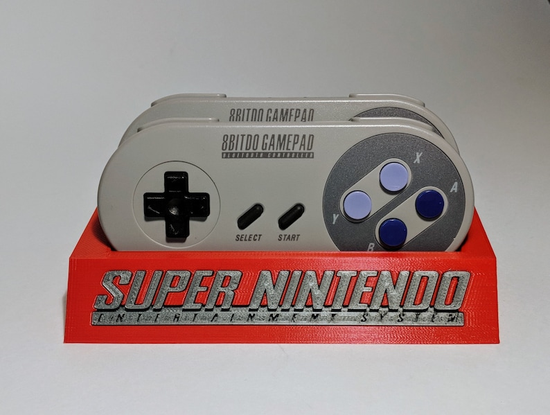 May include: A red plastic stand with the words "SUPER NINTENDO ENTERTAINMENT SYSTEM" printed on the front. Two gray 8BitDo Gamepad controllers are sitting on the stand.