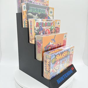 May include: A black, stepped display stand holding several Nintendo 64 video game boxes. The boxes are in protective cases and feature game titles like Star Fox and Super Smash Bros. The stand has the Nintendo 64 logo in blue.