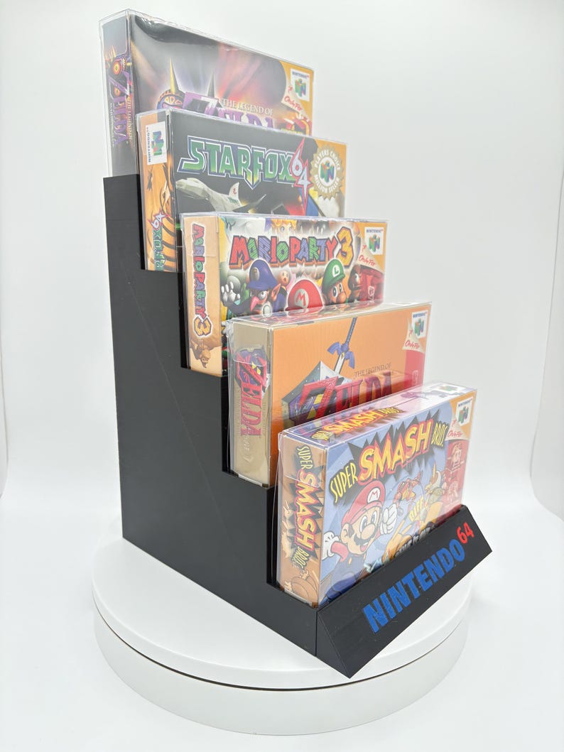May include: A black, stepped display stand holding several Nintendo 64 video game boxes. The boxes are in protective cases and feature game titles like Star Fox and Super Smash Bros. The stand has the Nintendo 64 logo in blue.