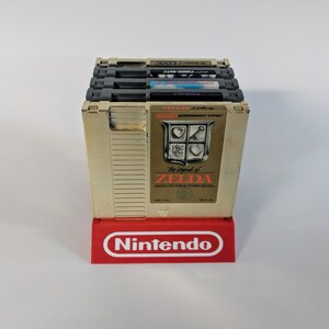 Op de afbeelding: Een stapel van vijf vintage Nintendo Entertainment System-spelcartridges, waaronder The Legend of Zelda, Super Mario Bros. 2, Punch-Out!!, en twee andere. De cartridges worden getoond op een rode Nintendo-standaard.