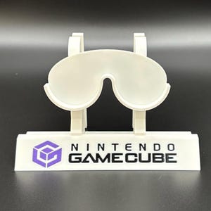 May include: A white plastic Nintendo GameCube controller stand. The stand features a stylized design with a purple GameCube logo and the words "NINTENDO GAMECUBE" in black. The stand is designed to hold a controller.